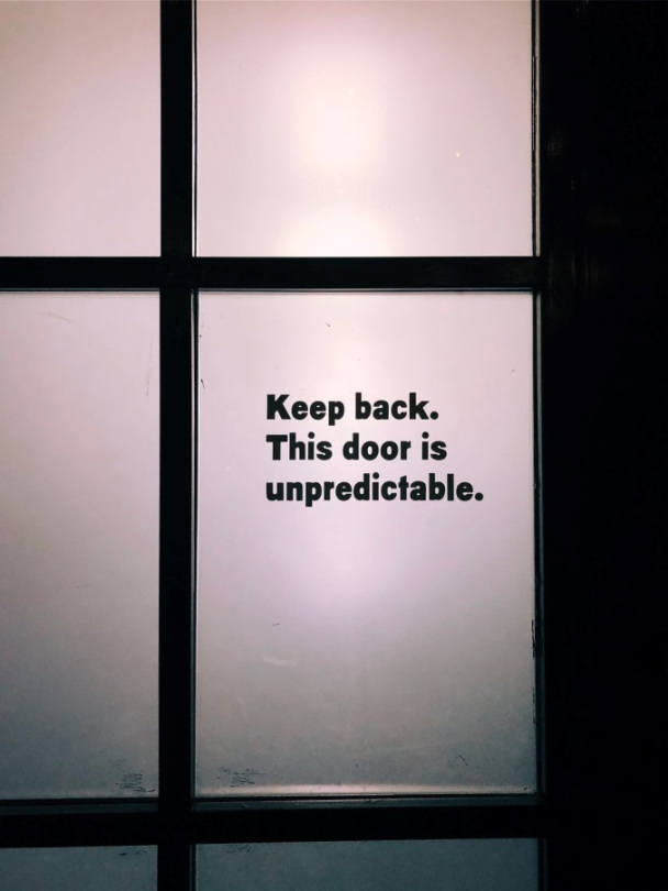 glass panel door with sign that says Keep Back. This door is unpredictable.